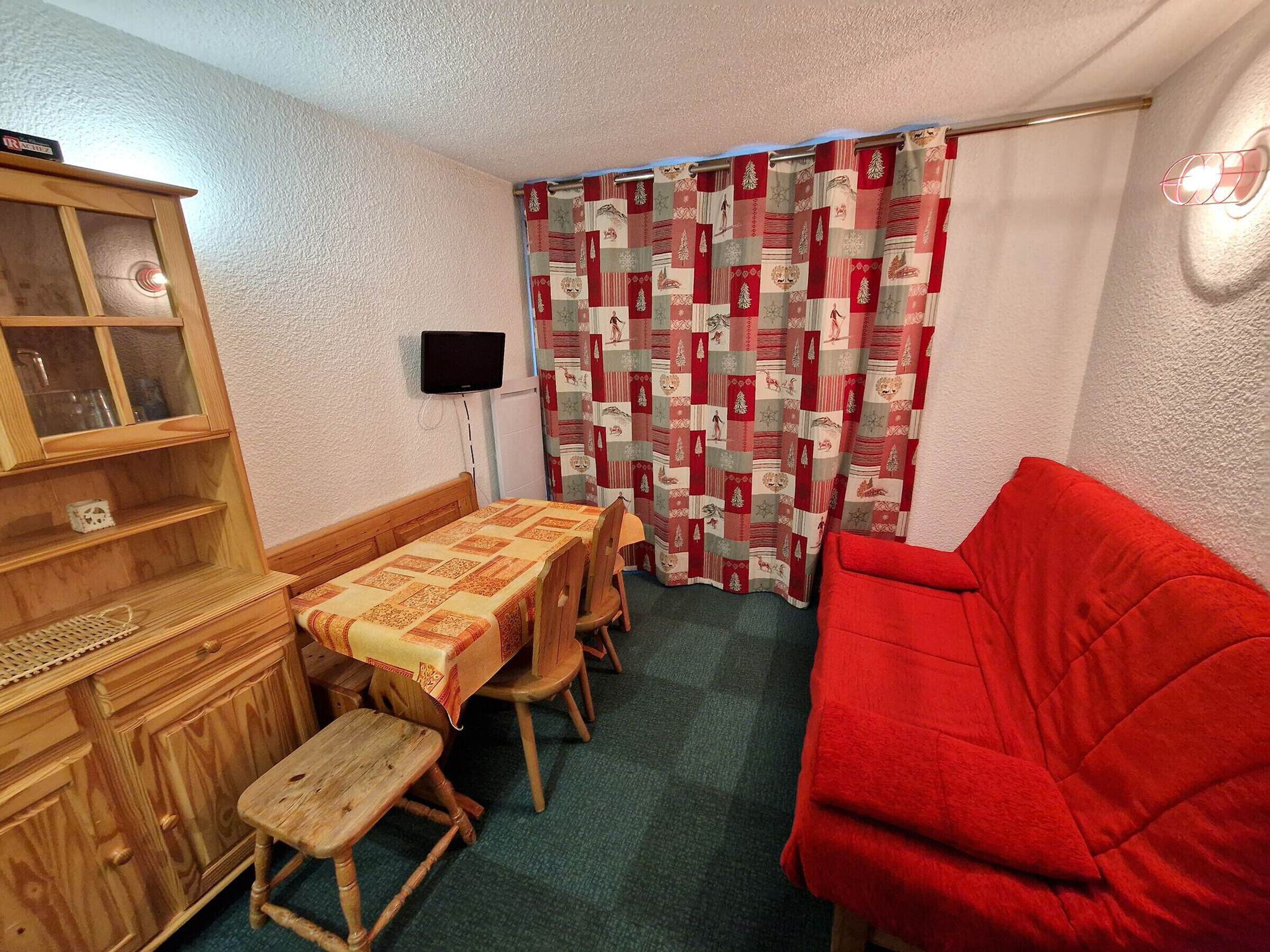Une petite pièce avec un meuble en bois, une table recouverte d’une nappe matelassée, un canapé rouge et un rideau à motifs.