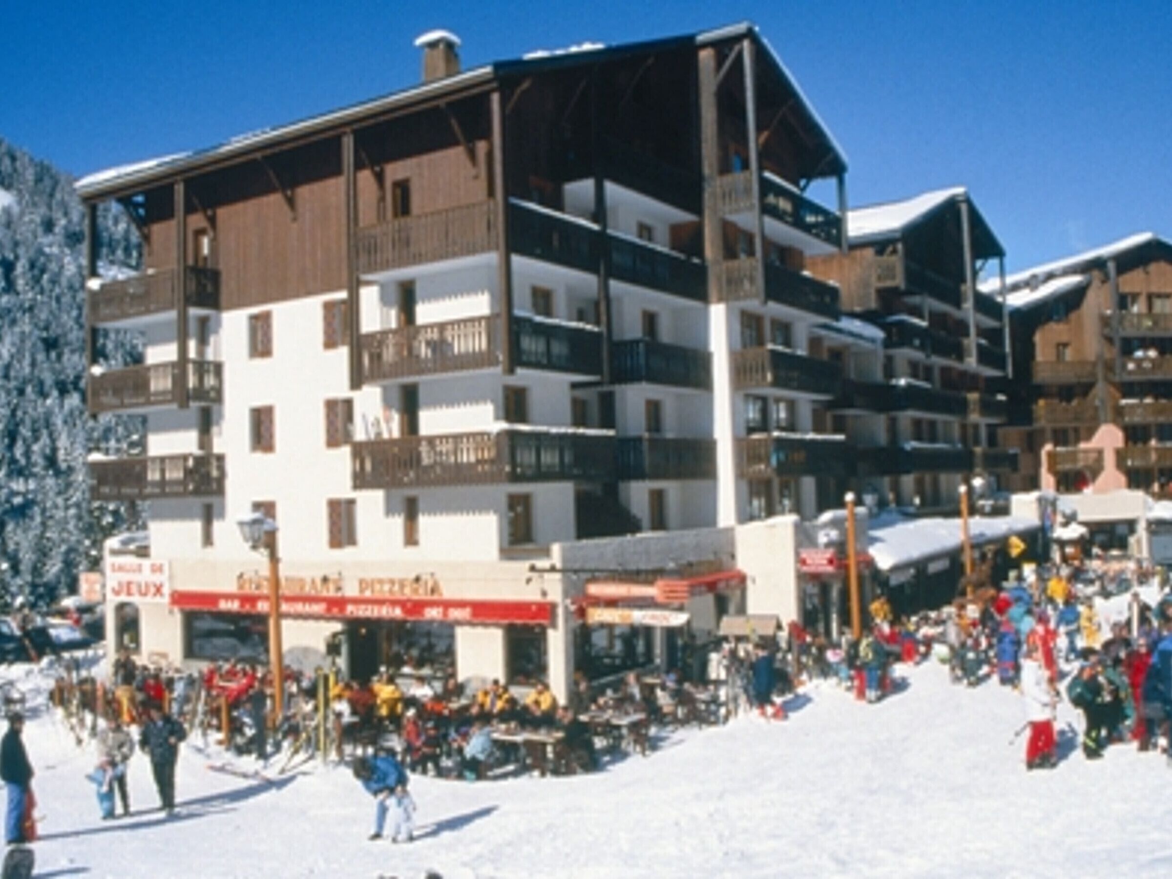 Een meerhoog gebouw met balkons, een skipiste op de voorgrond en een bord met daarop een restaurant.