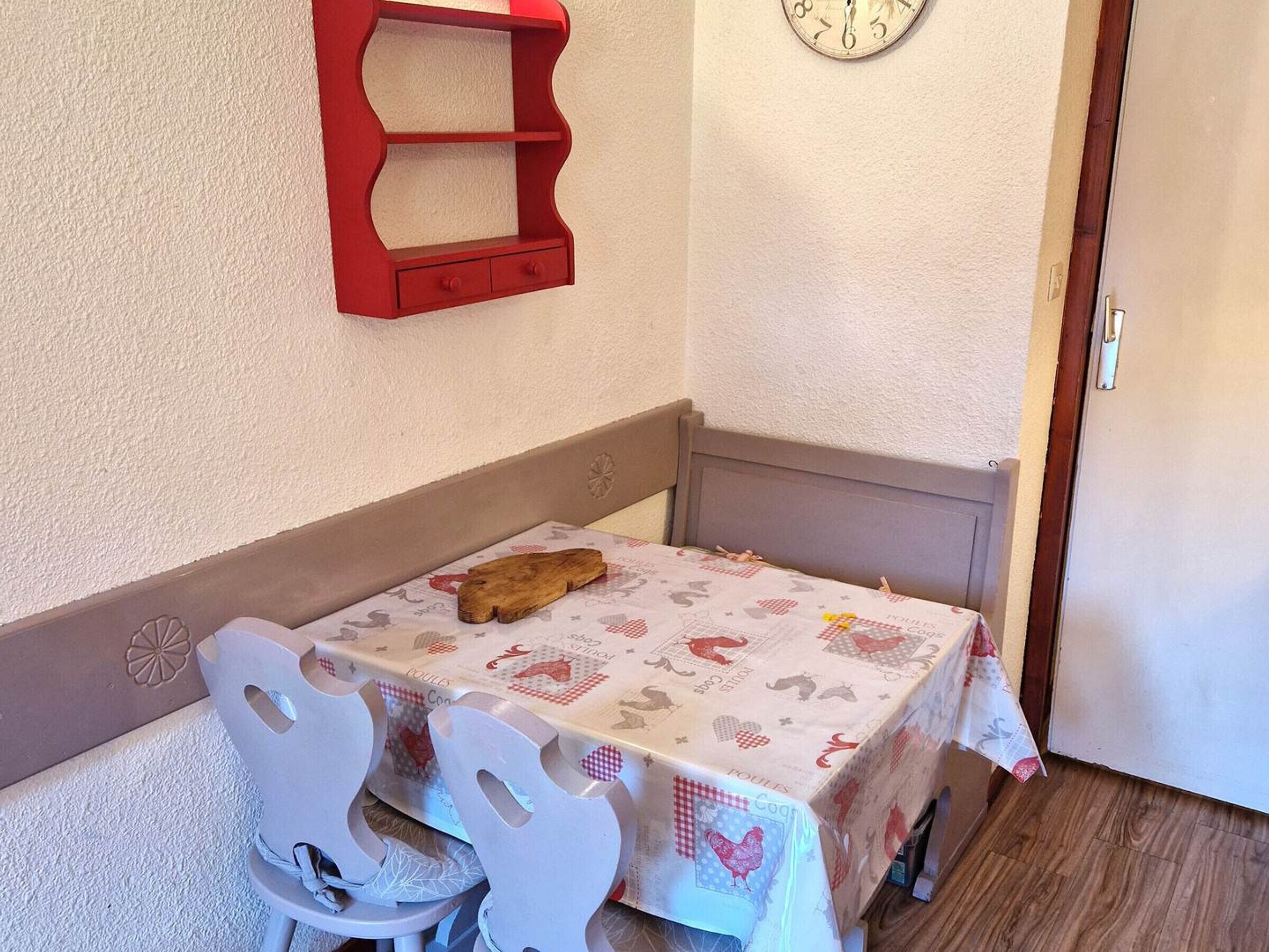 Un pequeño comedor con una mesa cubierta por un mantel estampado, un estante rojo fijado a la pared, un reloj de pared y una puerta al fondo.