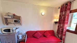 A compact room with a bed, a small desk with a microwave, a kettle, and a shelf with books and a plant.