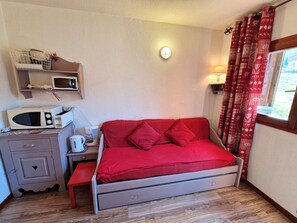 Habitación pequeña con cama, un escritorio con microondas, hervidor de agua y una estantería con libros y una planta.