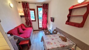 A compact room with a bed, a small table with a tablecloth, a bench, a wall-mounted shelf, and a window with curtains.