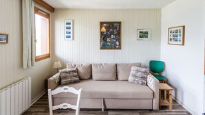 Ein Zimmer mit einer Couch, einem Sessel, einem kleinen Tisch, einem Fenster mit Vorhängen und Bildern an der Wand.