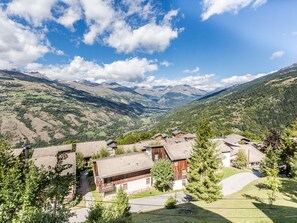 A mountain village with traditional wooden houses, a valley, and distant mountains under a partly cloudy sky.