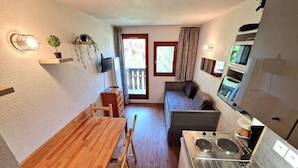 A compact kitchen with a wooden table and chairs, a built-in stove, and a small seating area with a sofa and curtains. - Renovated studio for 2-3 in Valfréjus, balcony and slope view (Valfréjus)