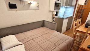 A bedroom with a bed, a bedside table, a chair, and a kitchenette in the background. - Renovated studio for 2-3 in Valfréjus, balcony and slope view (Valfréjus)