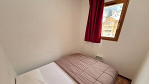 A small, single-bed room with a wooden floor, a window with a view of buildings, and a red curtain. - Valfréjus: 2-room + cabin, central location, 4/6 people (Valfréjus)