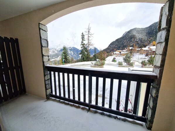 A balcony with a stone wall and a black railing overlooking a snowy mountain landscape with buildings.