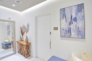 Una habitación moderna con un espejo grande, una mesa consola de madera con objetos decorativos, un banco azul y un cuadro en la pared.