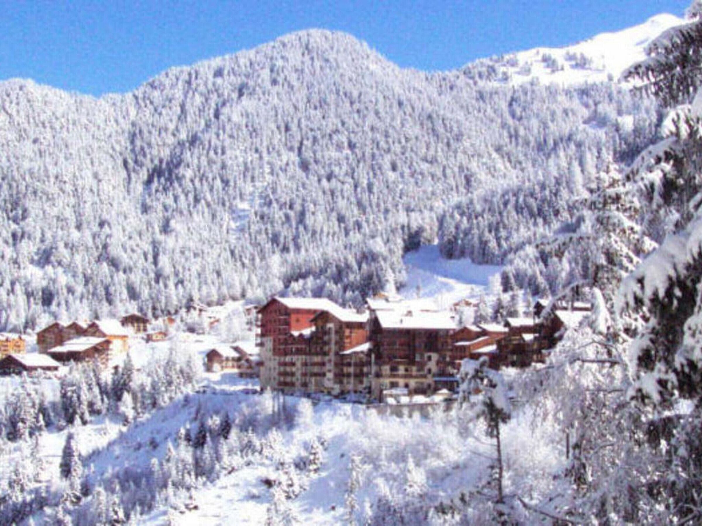 A snow-covered mountain landscape with a cluster of buildings, likely a ski resort, featuring multiple-story structures with red roofs.