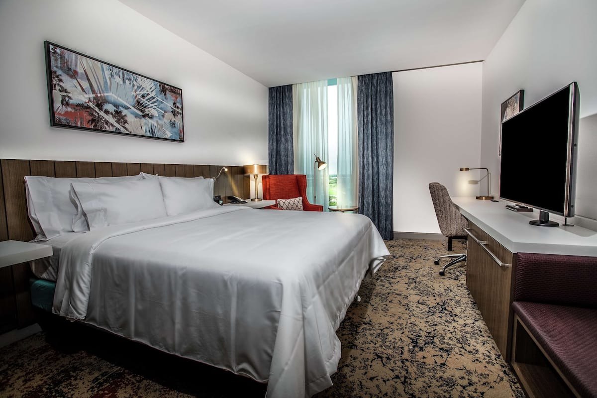 A hotel room with a large bed, a desk with a flat-screen TV, a chair, and a desk lamp.