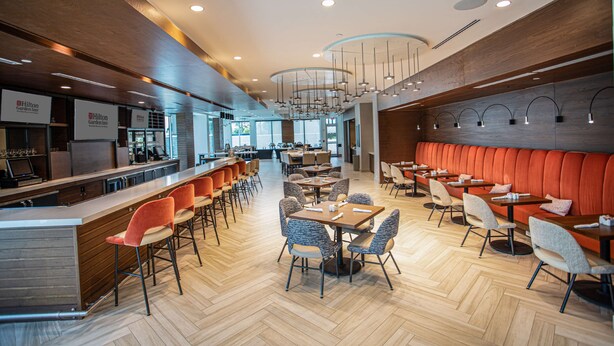 A modern hotel lobby with a bar area, seating arrangements, and a view of the kitchen in the background. - Hilton Garden Inn West Palm Beach I95 Outlets (West Palm Beach)