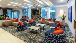 A modern lobby with sofas, armchairs, and a coffee table. There is a TV mounted on the wall, and potted plants are placed around the room. - Hilton Garden Inn West Palm Beach I95 Outlets (West Palm Beach)