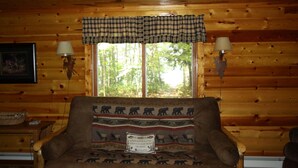 A cozy cabin interior with a wooden log wall, a plaid window valance, two wall-mounted lamps, and a sofa with a bear-themed throw pillow. - Peace, Privacy and a Pontoon!! No steps in home or to water! (Eagle River)
