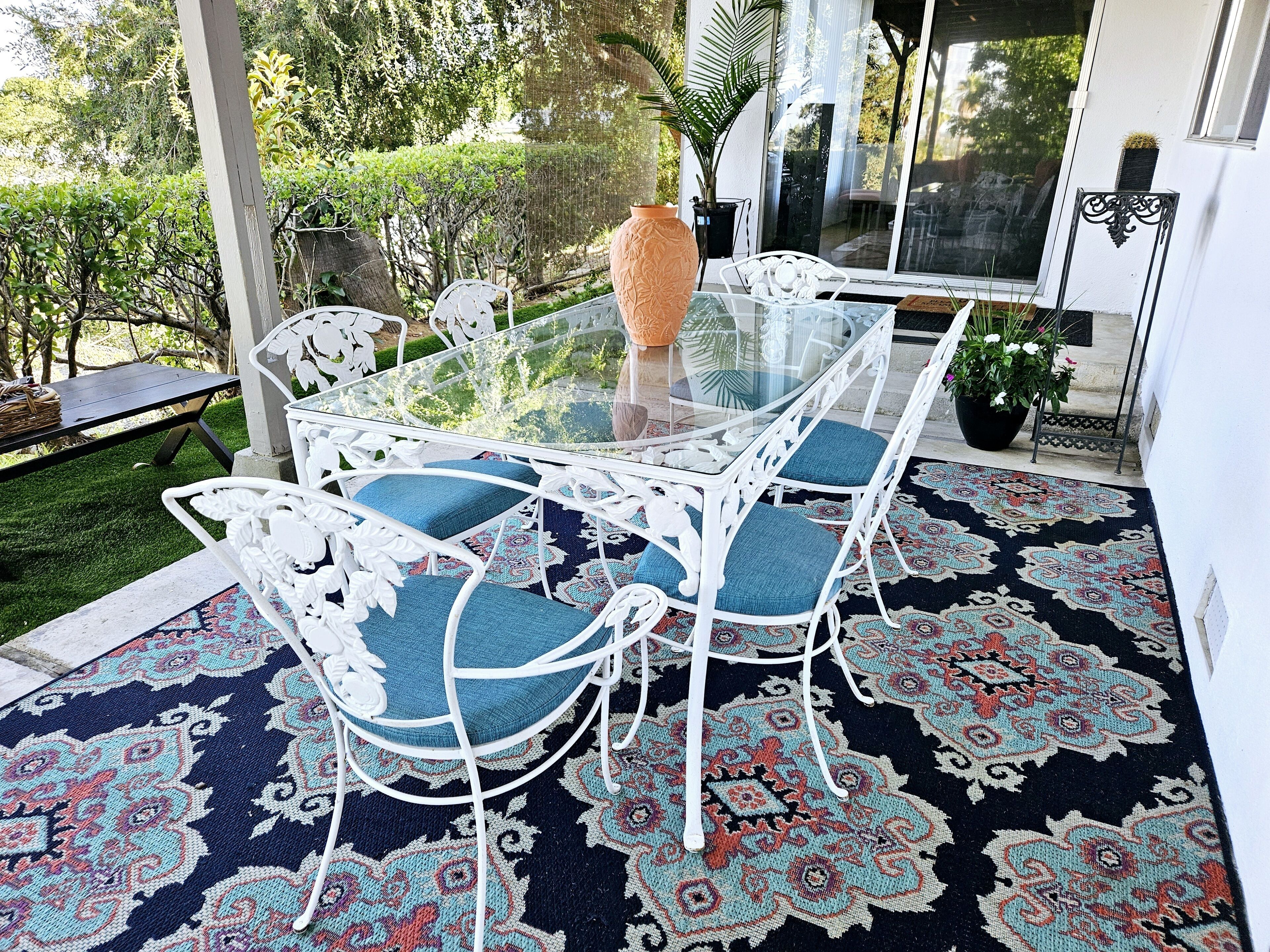 A glass-top table with white metal chairs, a patterned rug, and a potted plant.