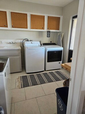 Compact kitchen with sink, washing machine, and dryer, featuring a corkboard and decorative sign.