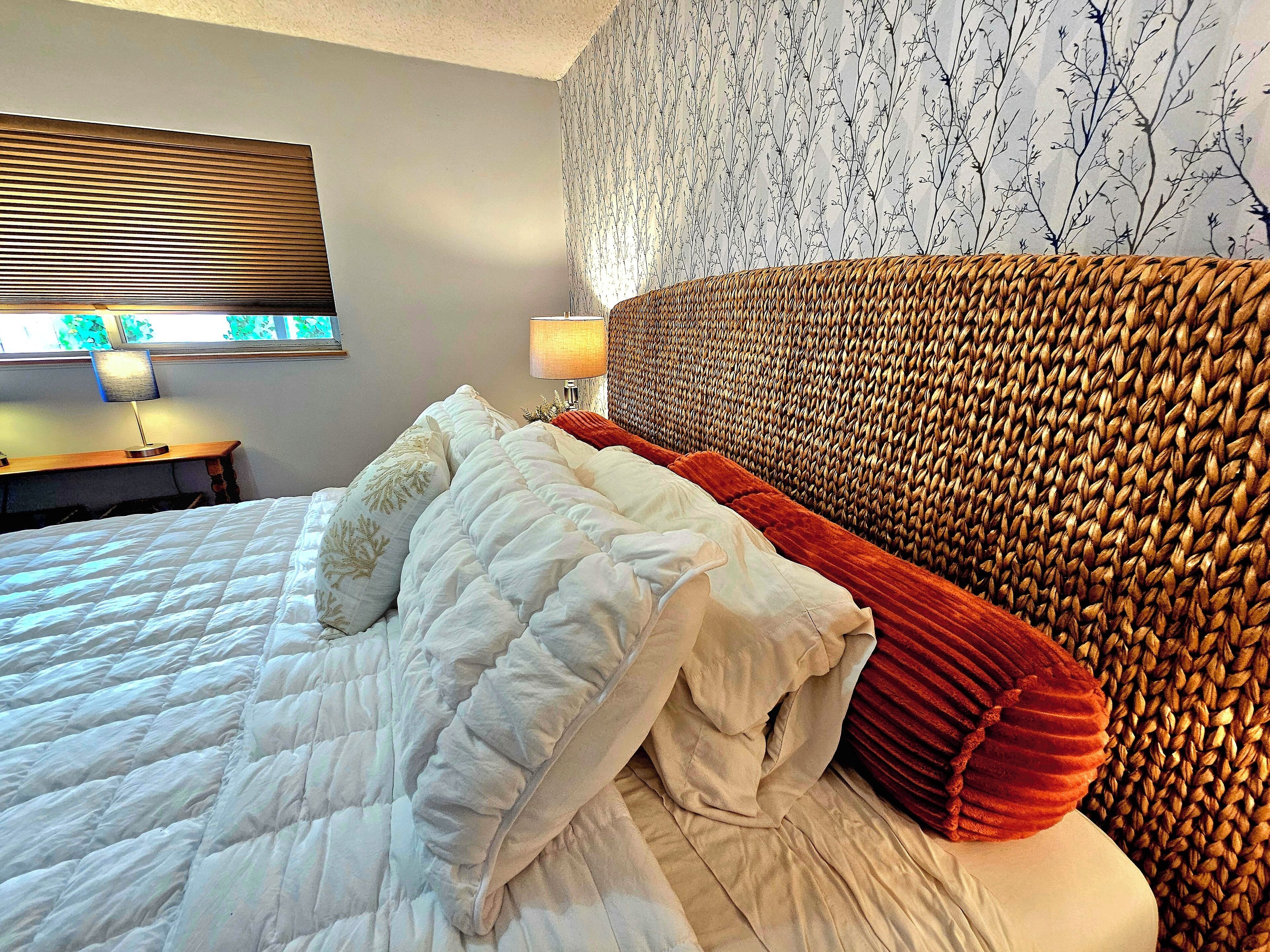 A neatly made bed with a white duvet and red pillows, a woven headboard, a bedside table with a lamp, and a wall with a tree branch pattern.