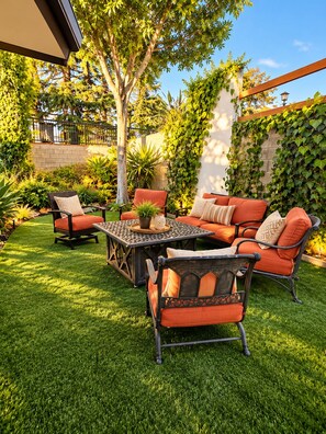 An outdoor patio with a seating area, including a sofa and chairs, surrounded by lush greenery and a brick wall.