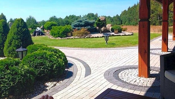 Ein gepflasterter Weg führt zu einer Laube mit Blick auf einen angelegten Garten und entfernte Gebäude.