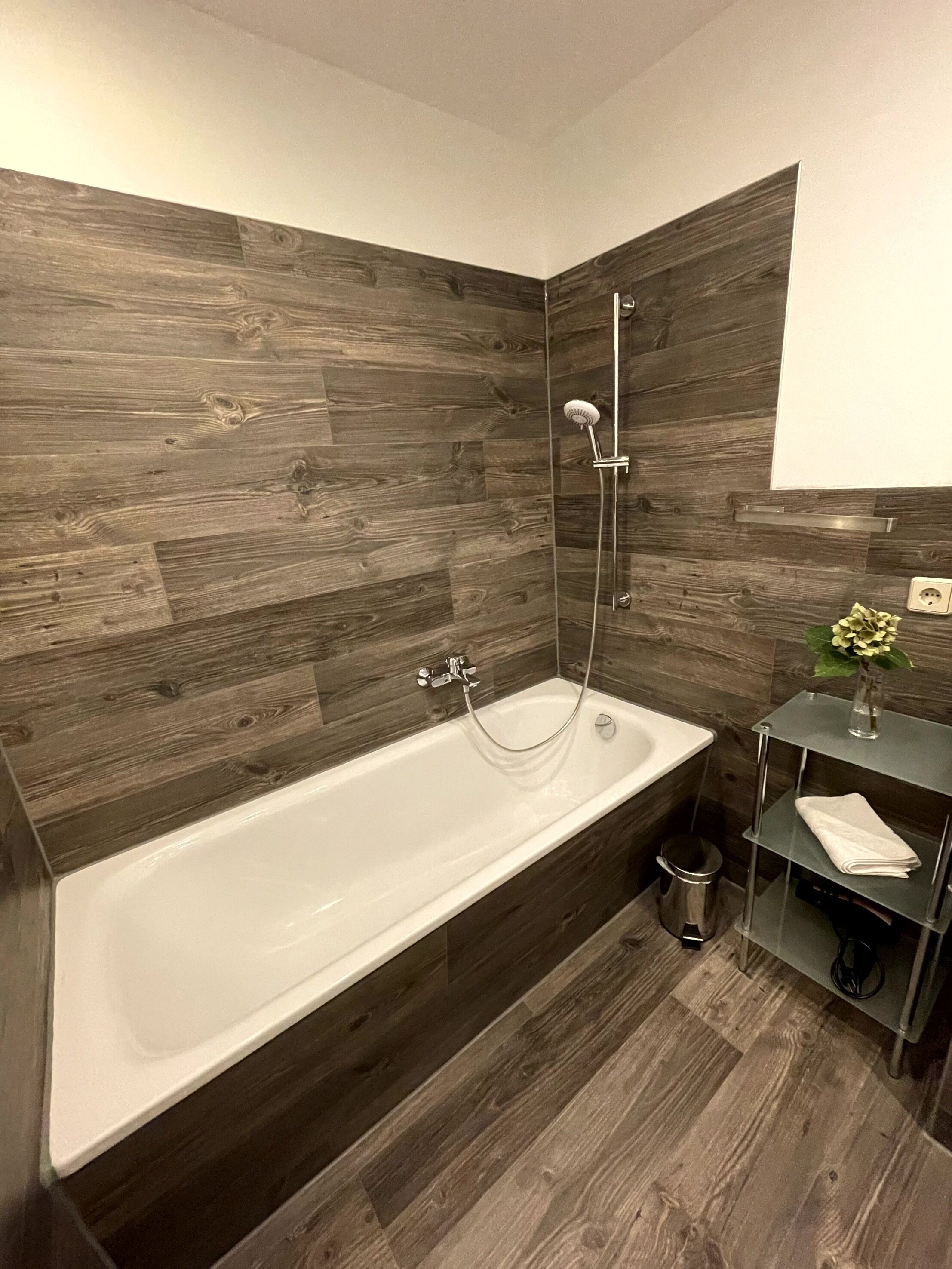 Un baño moderno con una bañera independiente, paneles de madera en la pared, un estante de vidrio con artículos de aseo y un pequeño cubo de basura.