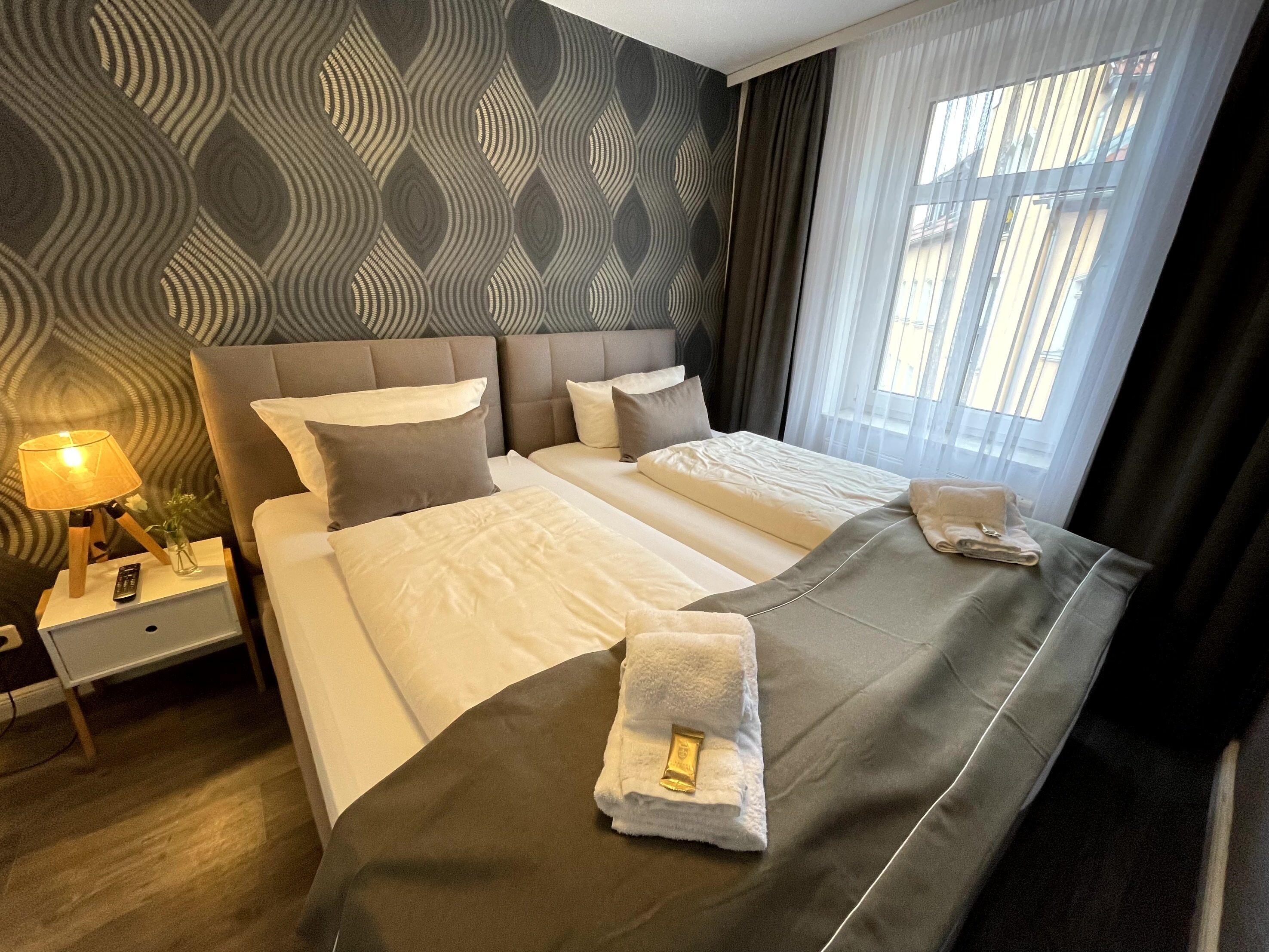 A hotel room with a large bed, a bedside table with a lamp, and a view of the outside through the window.