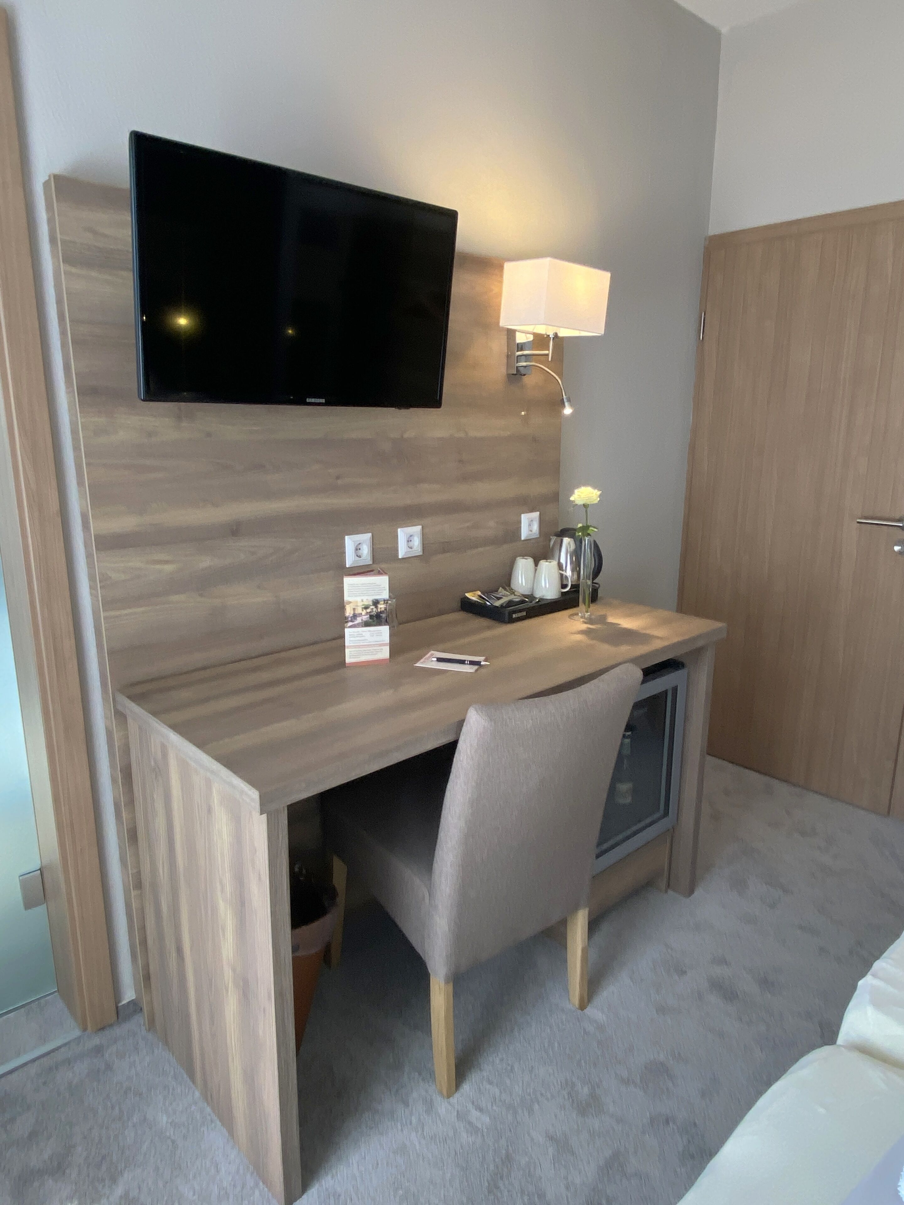 A compact hotel room with a wooden desk, a television mounted on the wall, a bedside lamp, and a wardrobe.