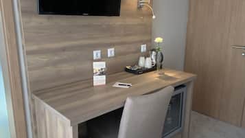 A compact hotel room with a wooden desk, a television mounted on the wall, a bedside lamp, and a wardrobe.