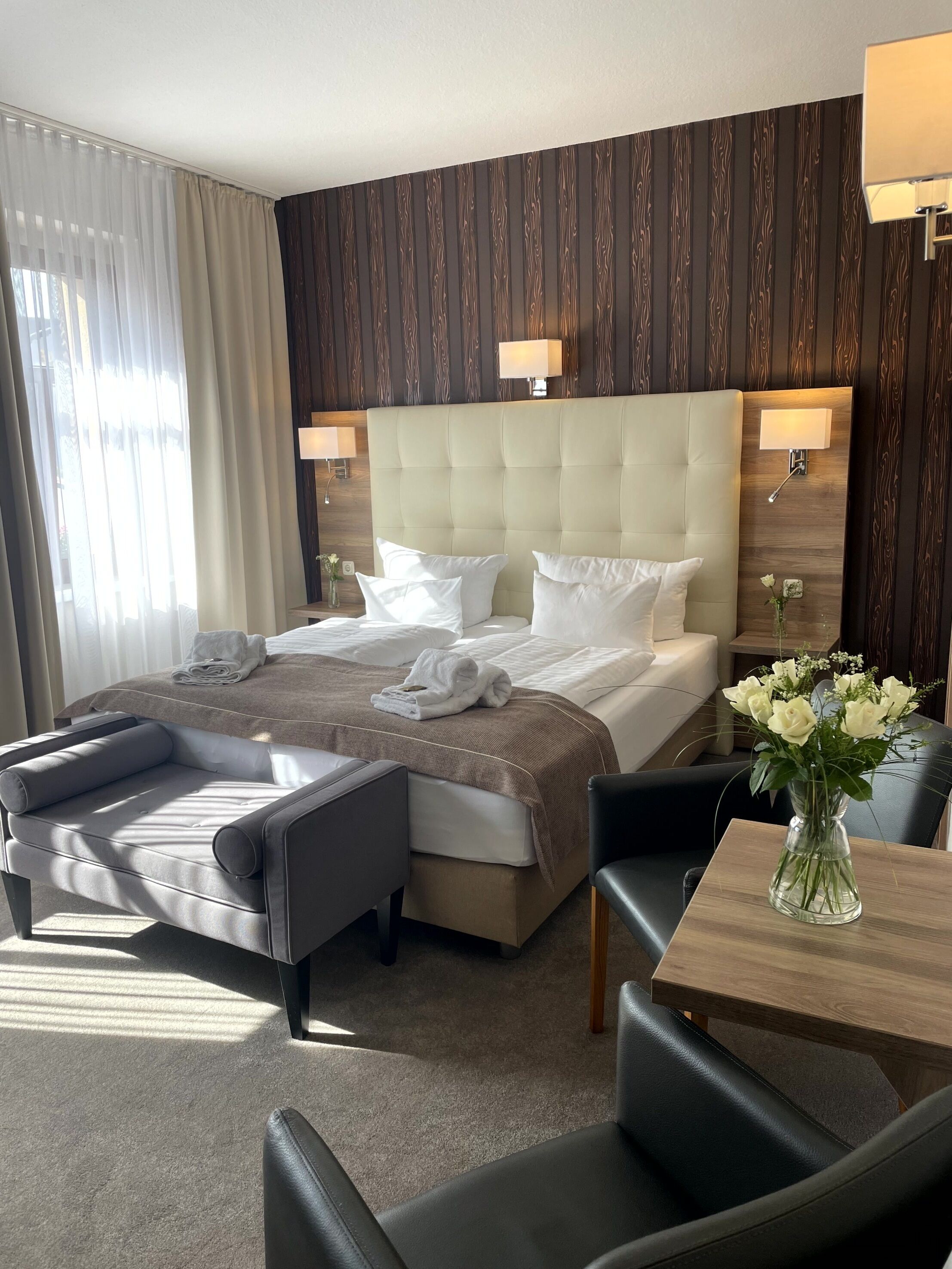 Habitación de hotel con una cama grande, dos mesitas de noche con lámparas, un sofá, una pequeña mesa con un florero y vistas al exterior a través de cortinas translúcidas.