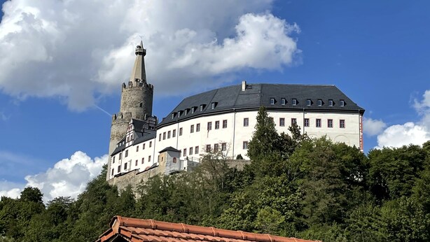 Een historisch kasteel met een opvallende toren en meerdere gebouwen, omgeven door weelderige begroeiing.