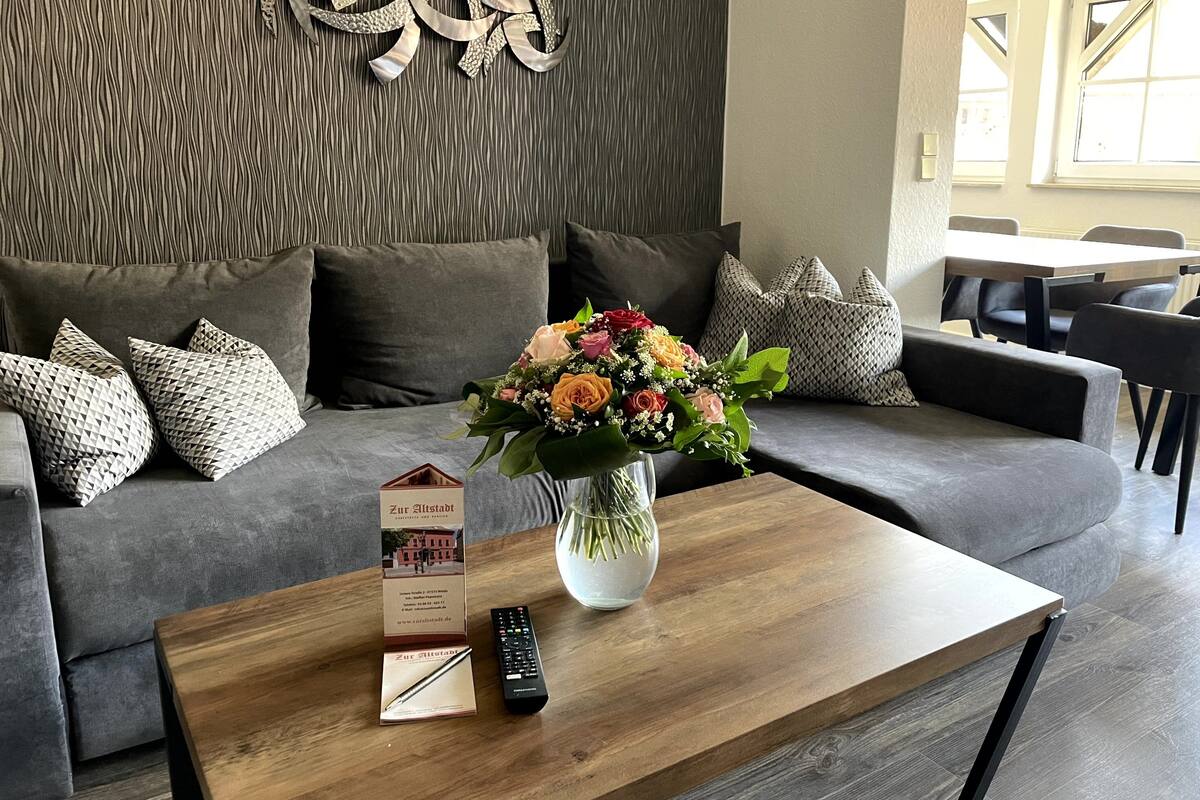 A modern living room with a grey sofa, patterned cushions, a wooden coffee table, a vase with flowers, a remote control, and a brochure.
