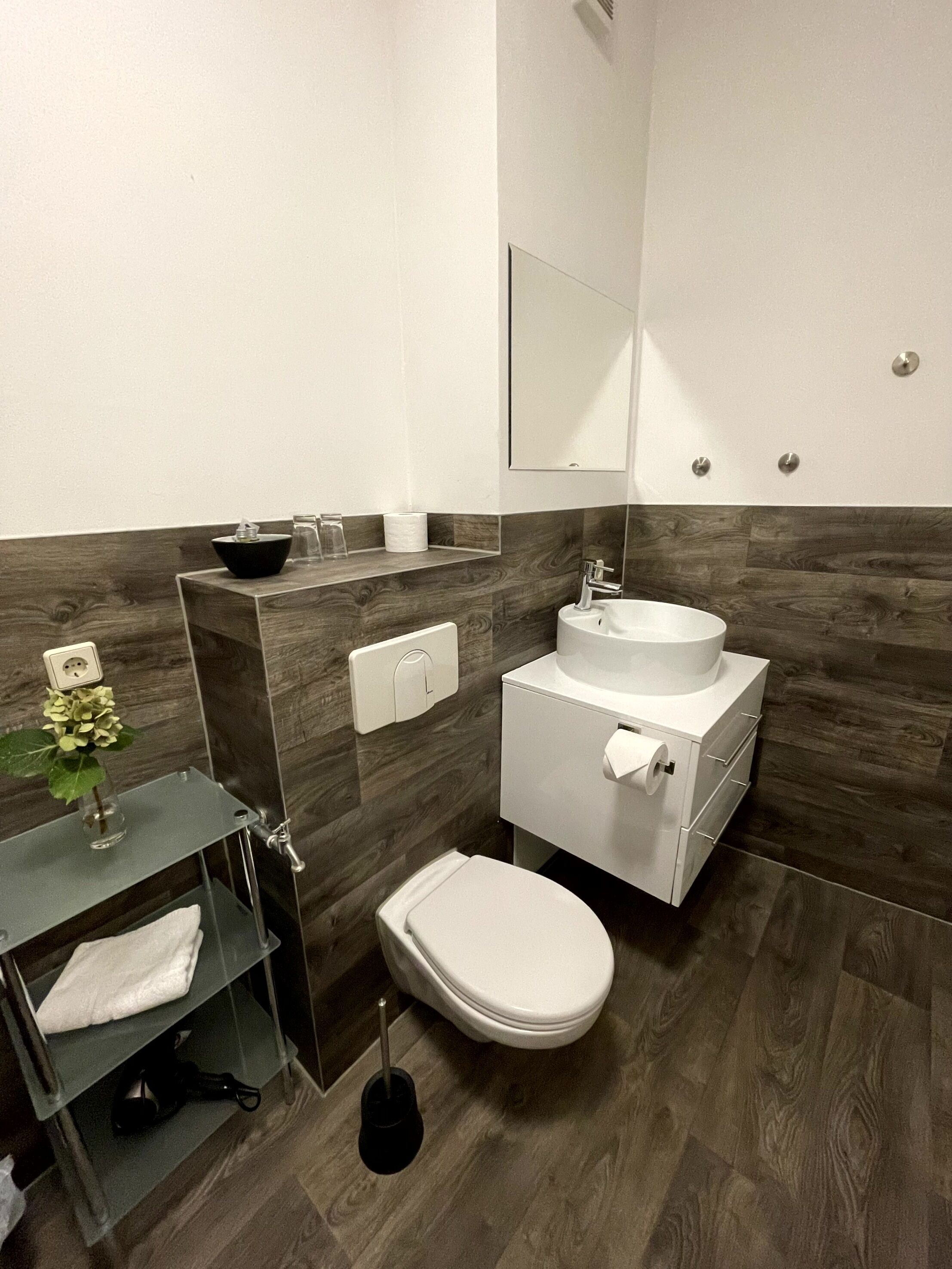 Ein modernes Badezimmer mit Toilette, Waschbecken und Spiegel. Ein Regal zeigt eine Vase und eine Flasche.