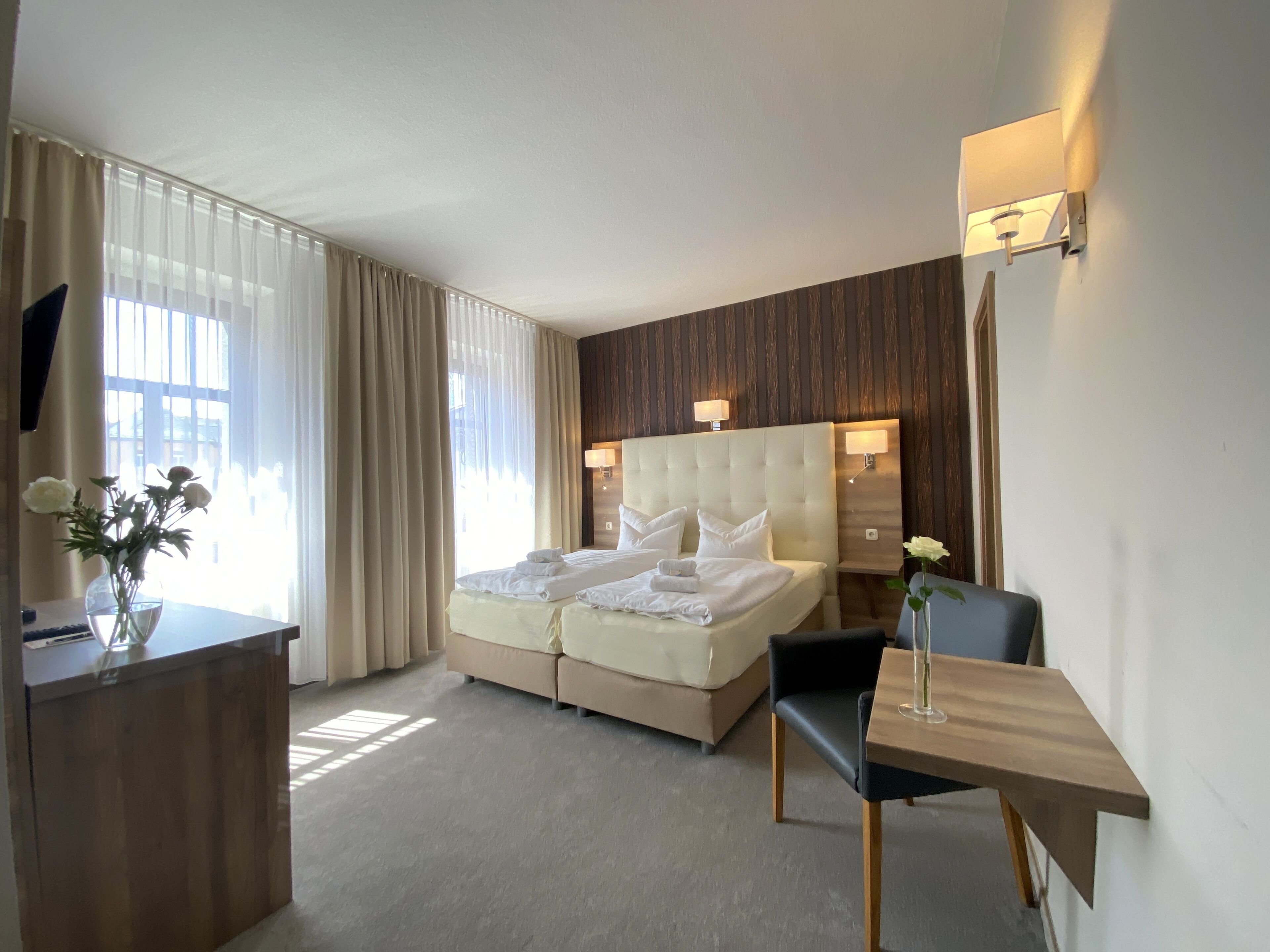 Ein Hotelzimmer mit Bett, Schreibtisch, Stuhl, Blumen in einer Vase und einem Fenster mit Vorhängen.