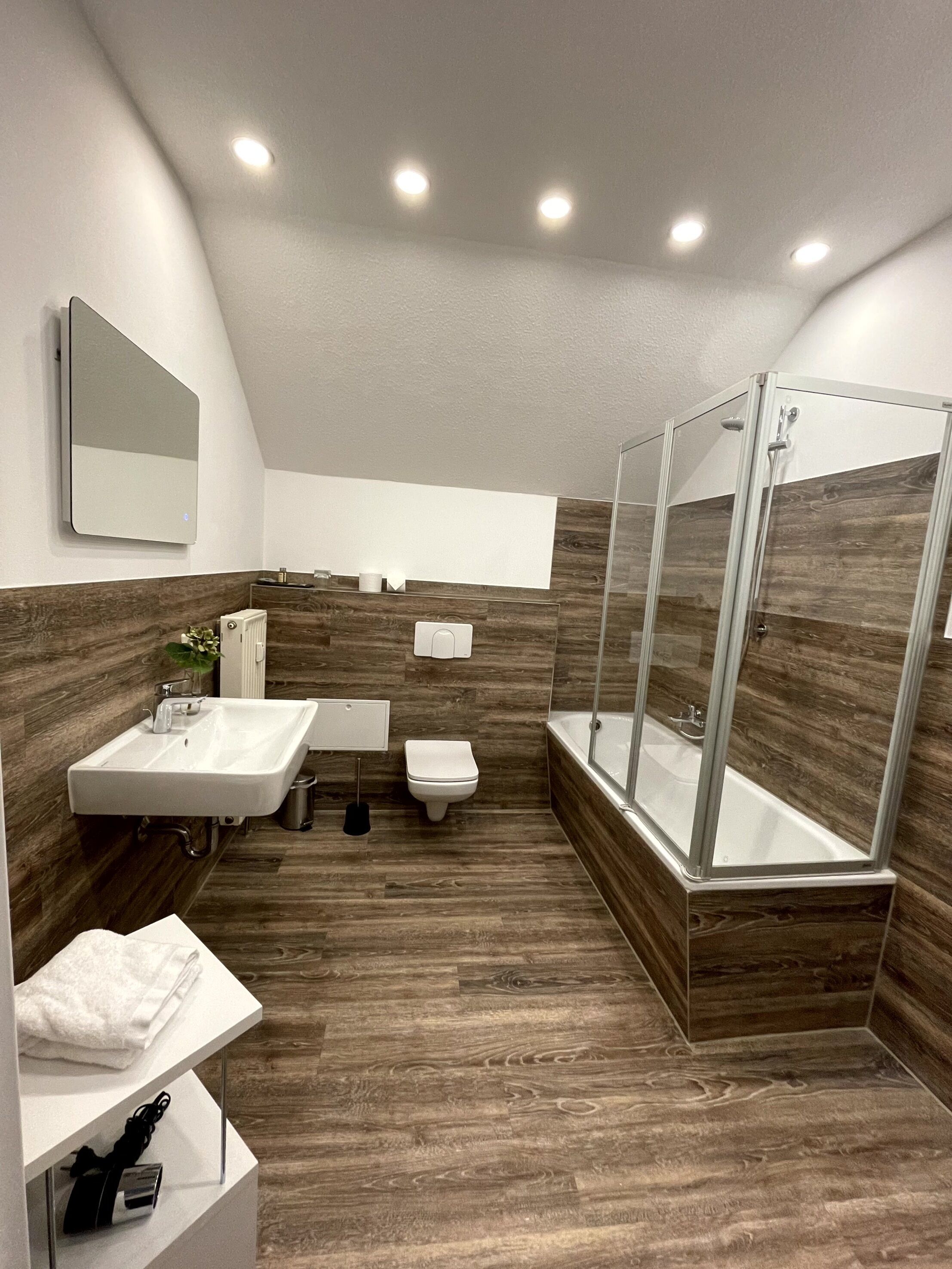 Un baño moderno con suelo de madera, ducha con cerramiento de vidrio, lavabo blanco y retrete.