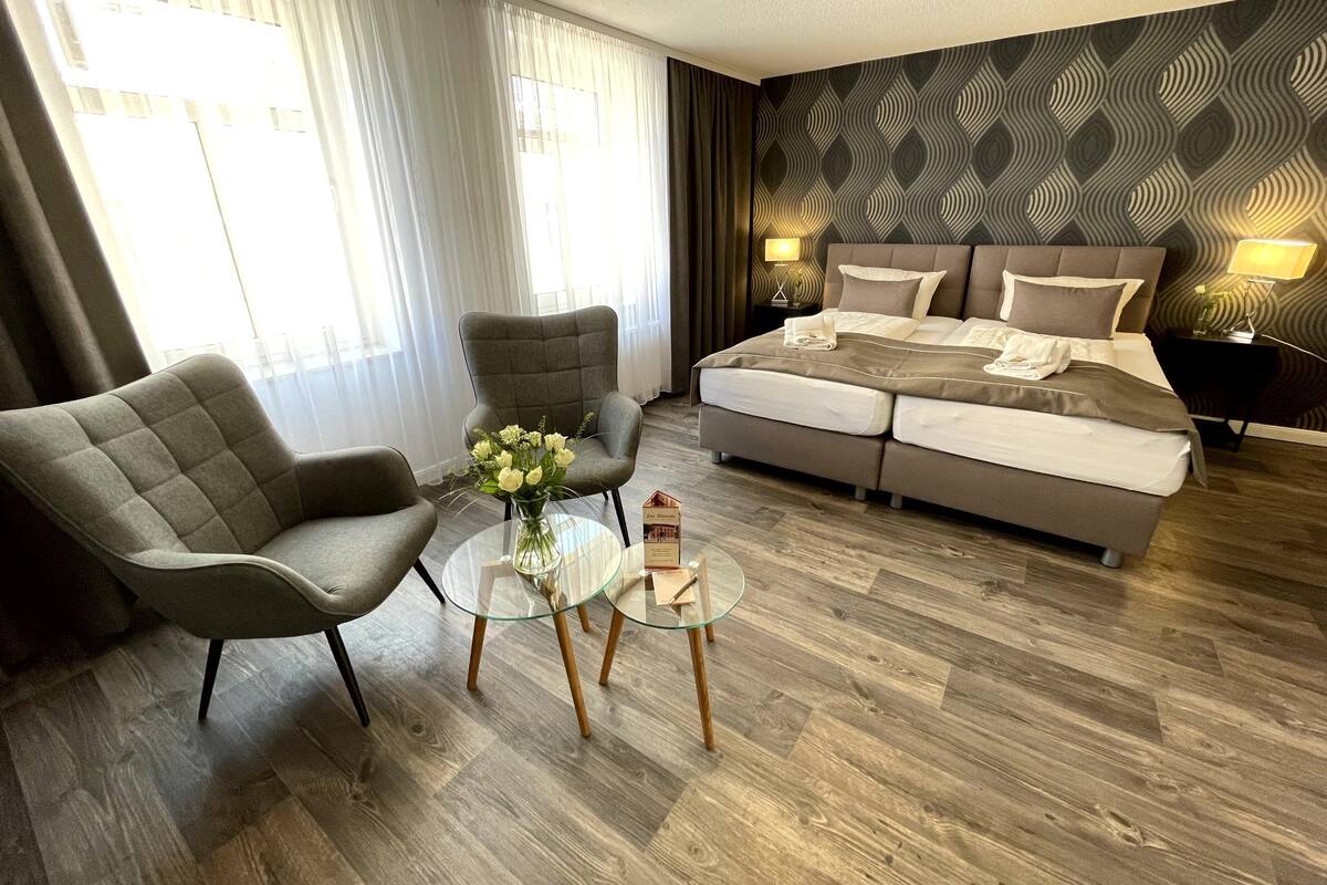 A modern hotel room with a large bed, two armchairs, a glass coffee table, and a patterned wall.