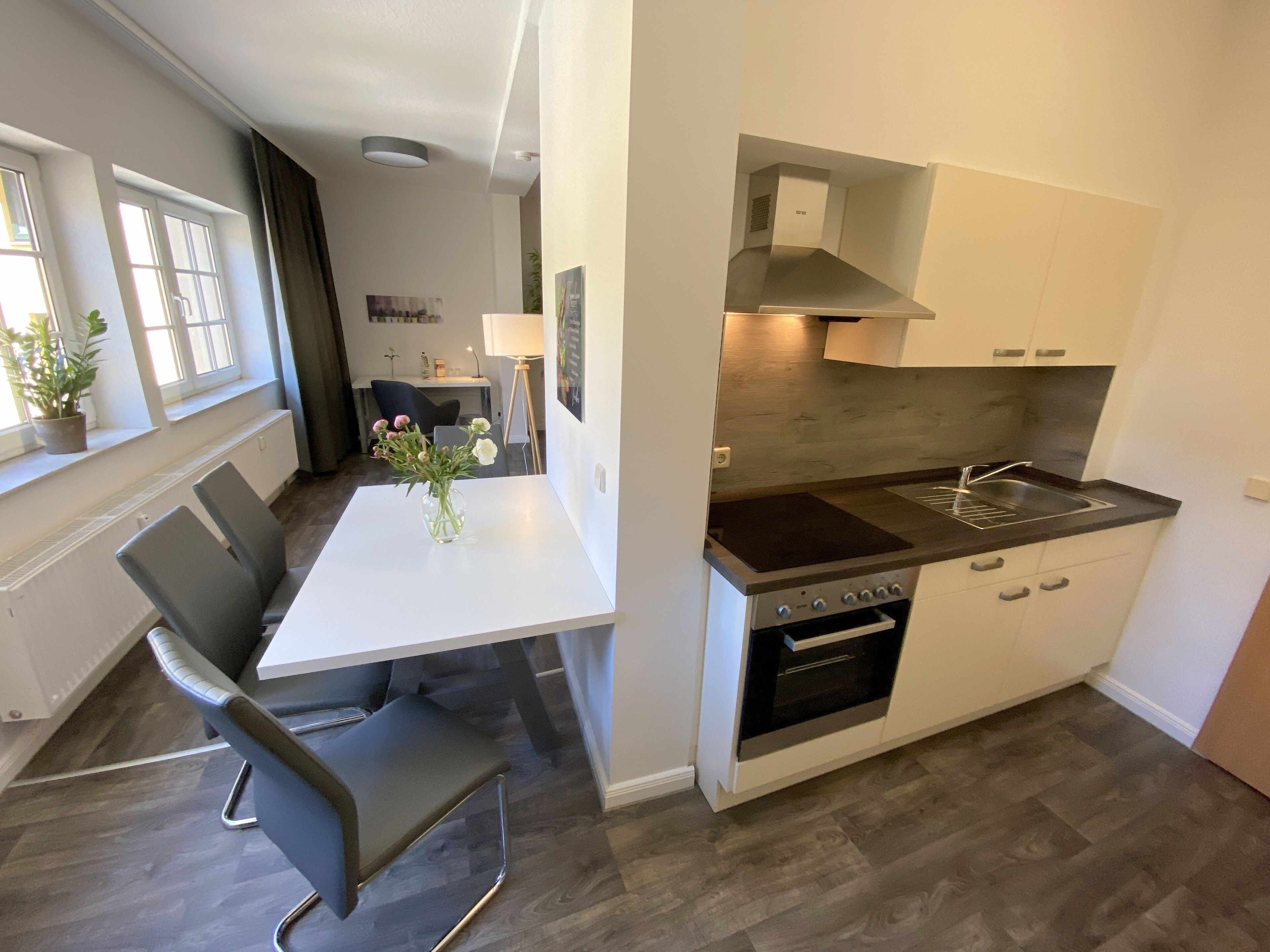 A modern kitchen with white cabinets, a built-in oven, and a dark countertop. There is a dining table with chairs and a vase with flowers on it.