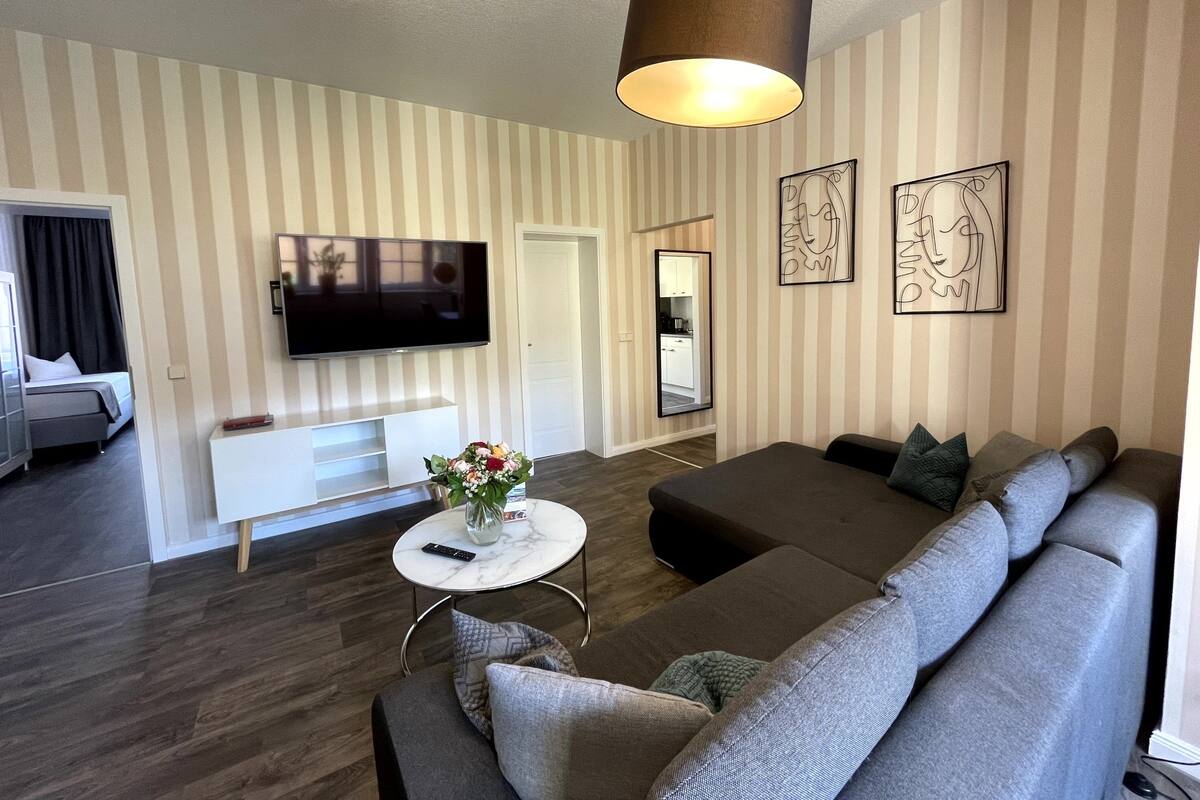 A modern living room with a large grey sofa, a small white coffee table, a TV mounted on the wall, and a striped wallpaper design.