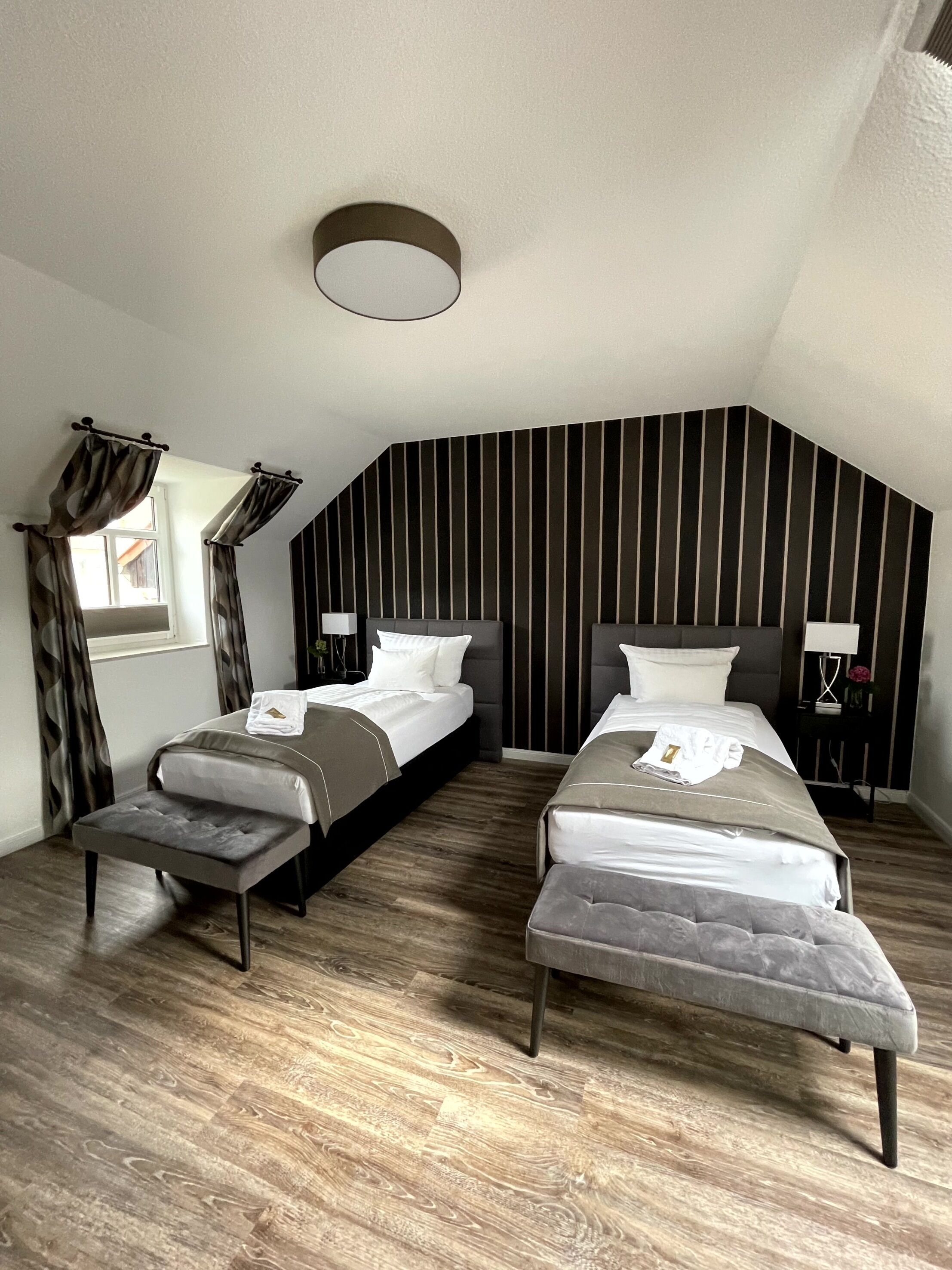A modern bedroom with two beds, a bench, a window with curtains, a wall-mounted light, and a striped wall design.