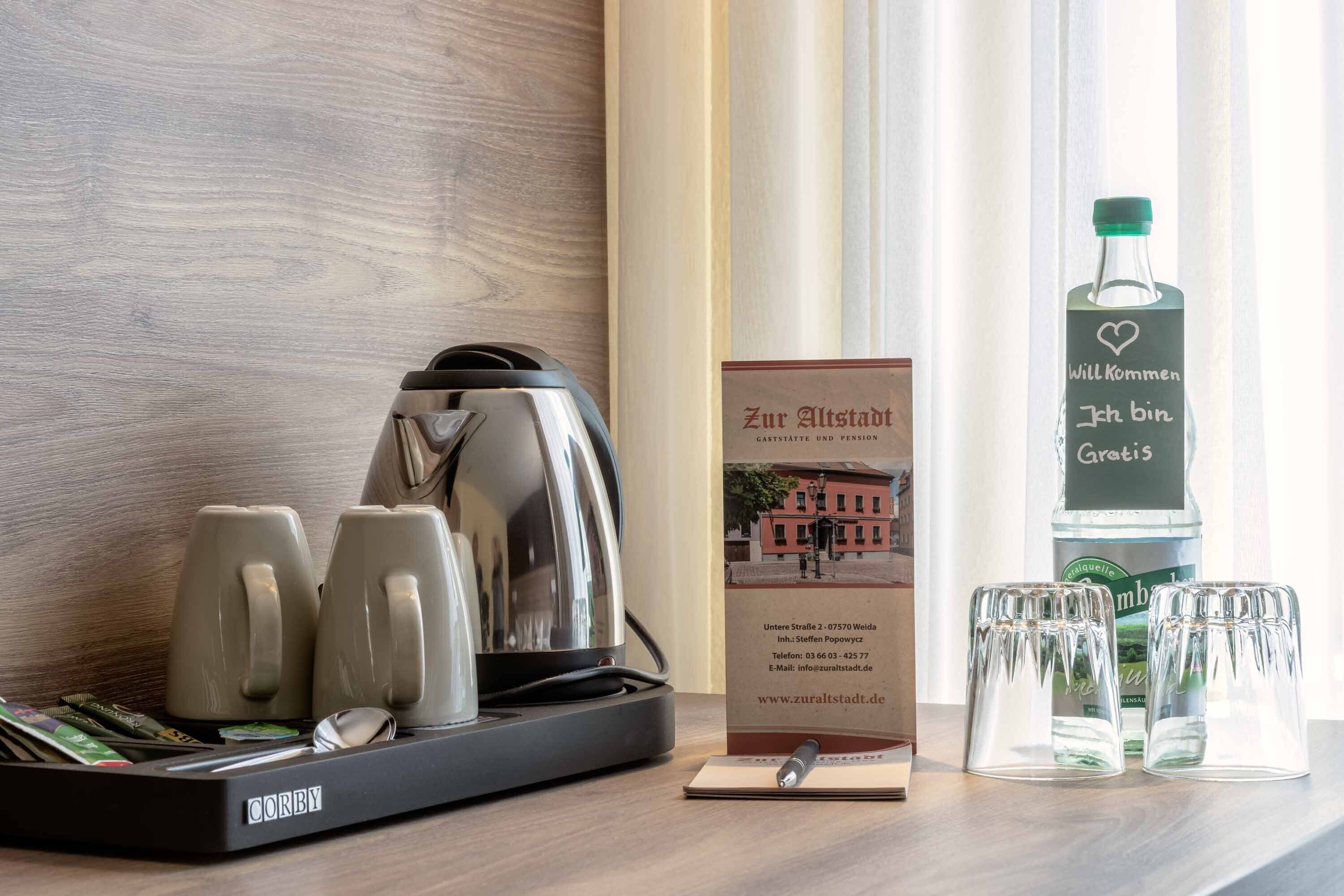 A hotel room with a coffee maker, two mugs, a spoon, a glass carafe, and a welcome card on the table.