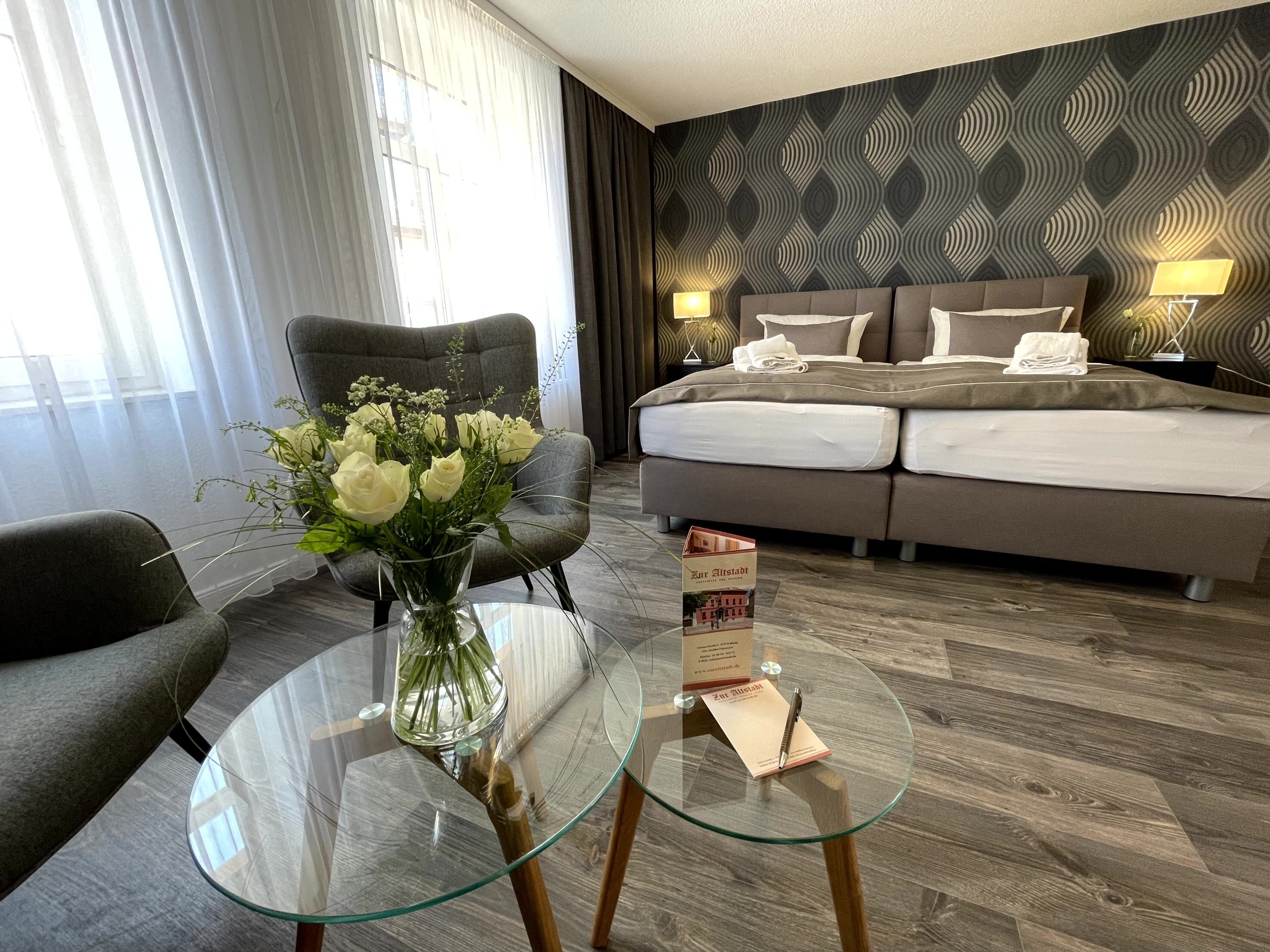 Una habitación de hotel con una cama, una silla, una mesita de vidrio y un jarrón con flores.