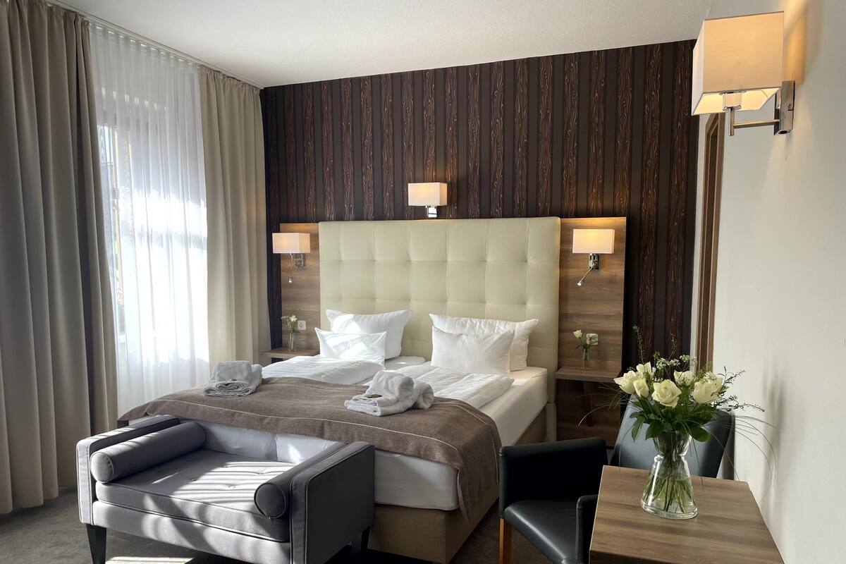 A hotel room with a large bed, a sofa, a small table, and a chair. There are two wall-mounted lamps and a vase with flowers on the nightstand.
