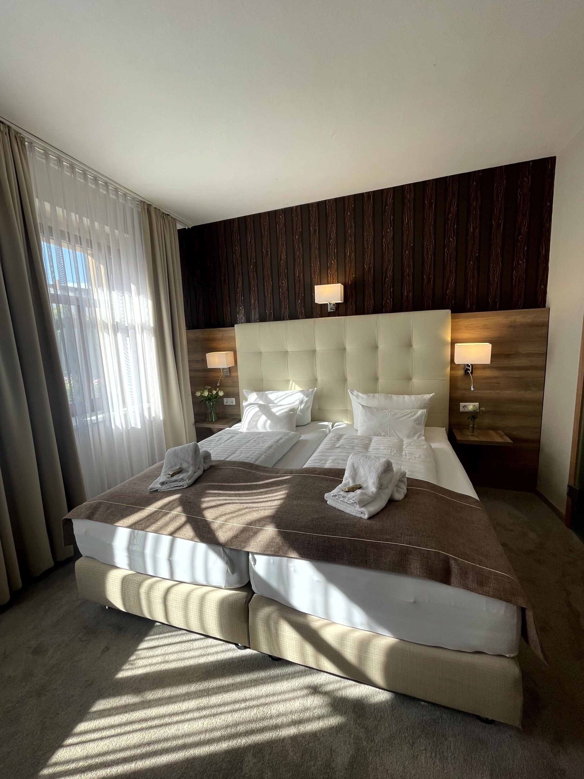 Une chambre d’hôtel avec un grand lit, deux tables de chevet avec des lampes, une tête de lit en bois et des rideaux légers.