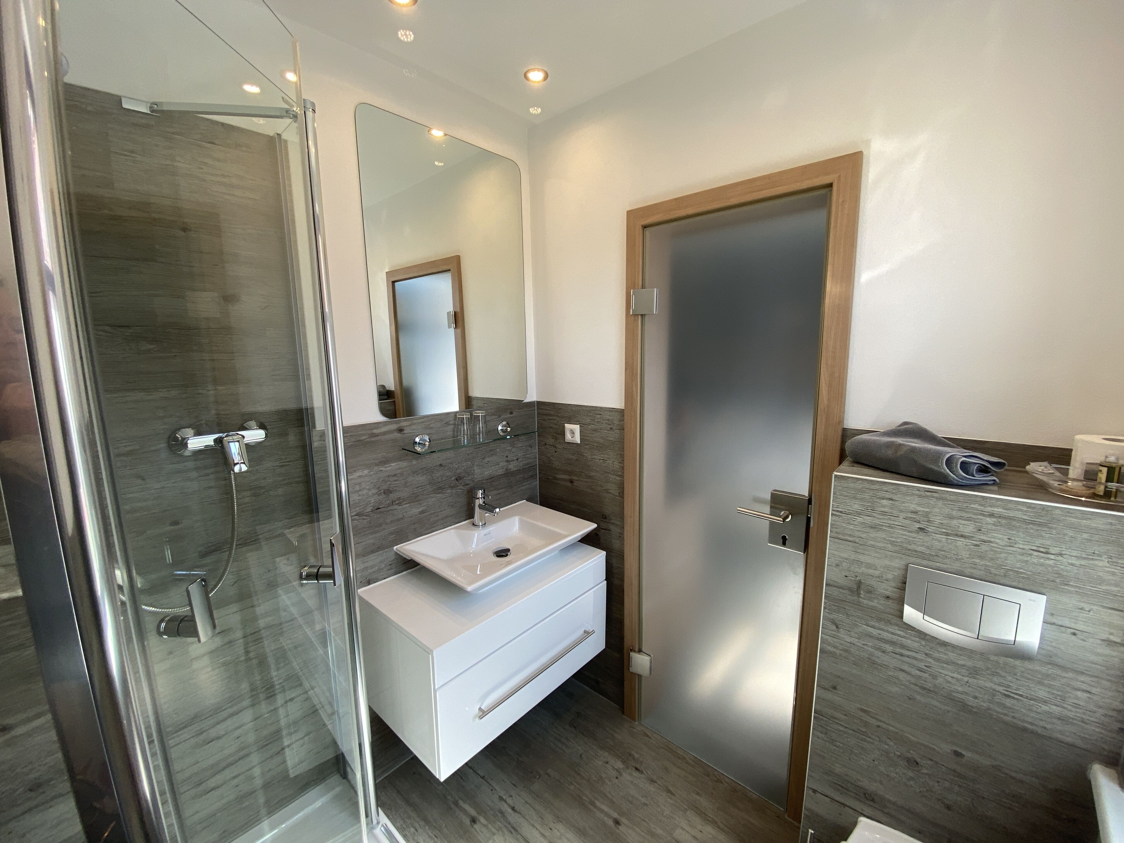 Un baño moderno con cabina de ducha de vidrio, tocador blanco con lavamanos y puerta de madera.