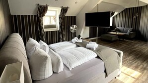 Een ruime slaapkamer met een groot bed, een flatscreen televisie, een zithoek met een bank en fauteuils, en een houten vloer.