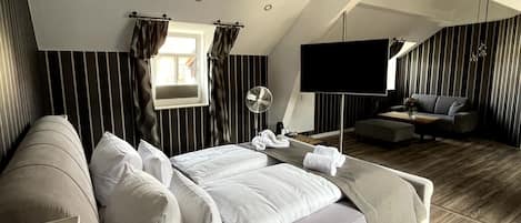 Un dormitorio amplio con una cama grande, un televisor de pantalla plana, una zona de estar con sofá y sillones, y piso de madera.