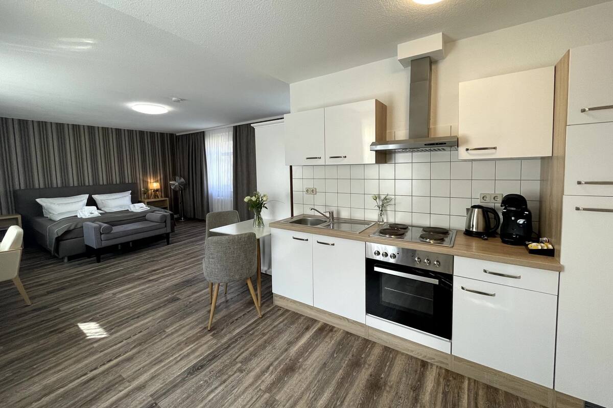 A modern hotel room with a kitchenette, a sofa, a dining table, and a large window with curtains.