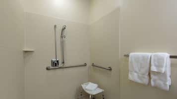 Un baño con paredes de azulejos blancos, una ducha con barras de apoyo, una silla blanca y un perchero con toallas.