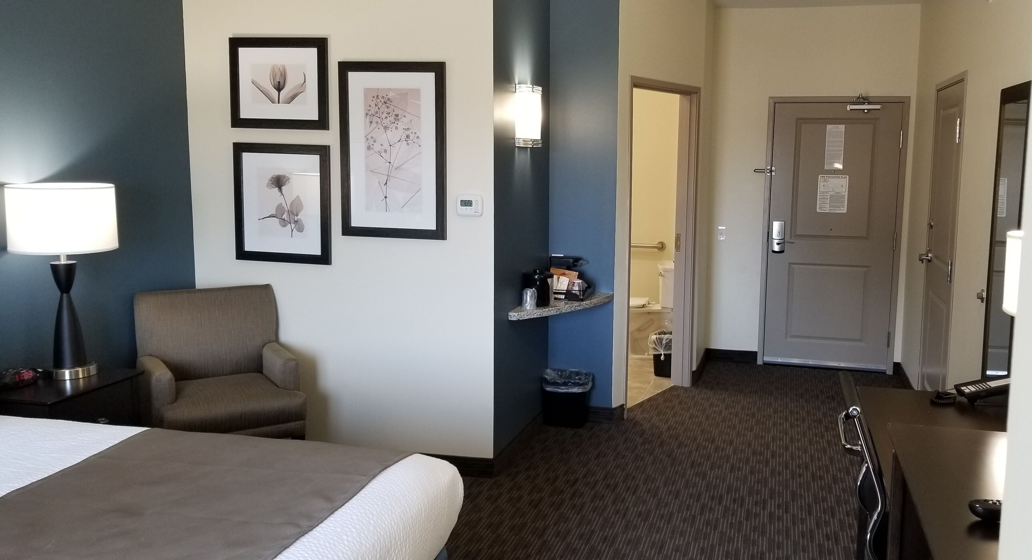 A hotel room with a bed, a chair, a desk with a lamp, framed pictures on the wall, a door, and a trash bin.