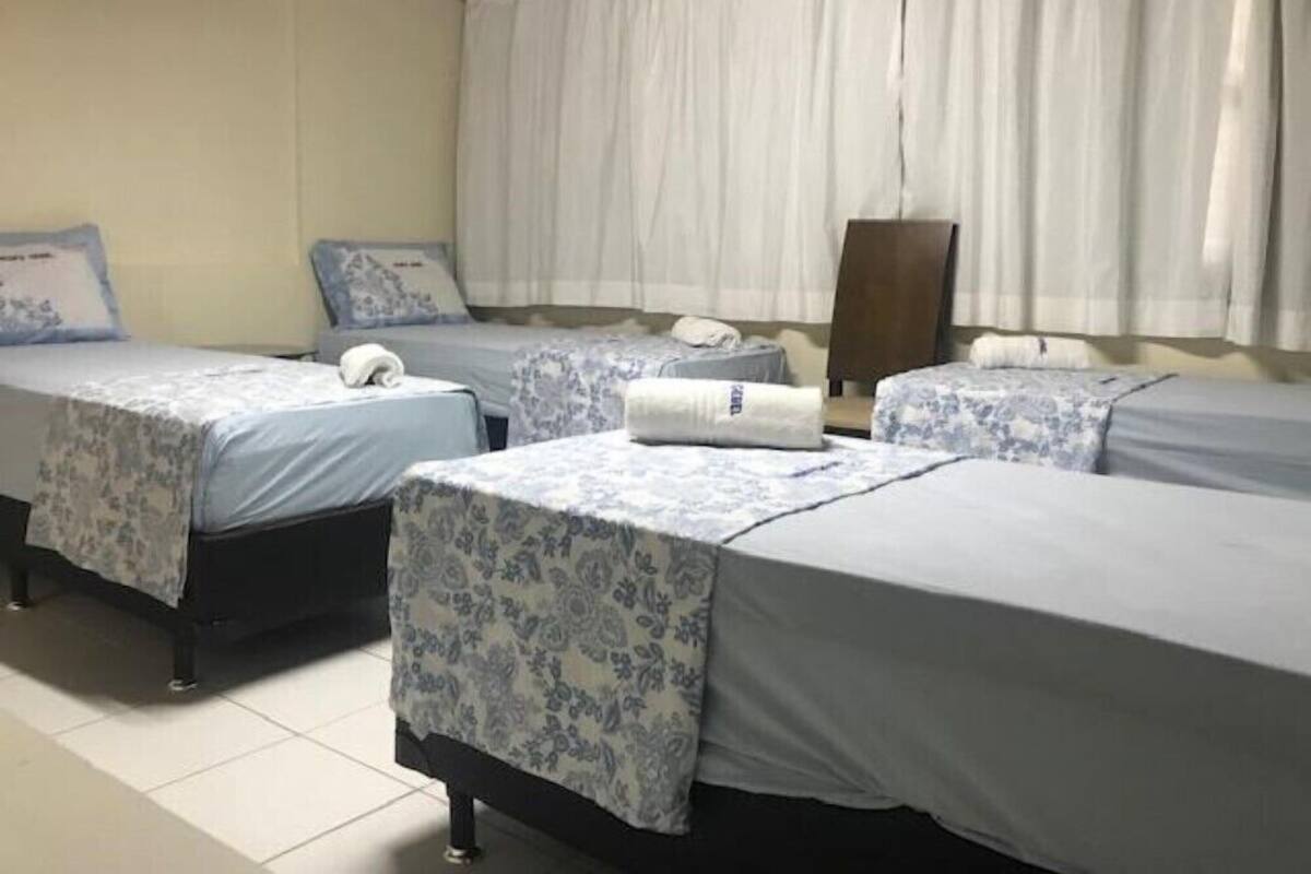 Quarto de hotel com três camas de solteiro, cada uma com colcha e almofadas estampadas com motivos florais. O quarto possui uma janela grande com cortinas brancas, teto de madeira e piso de azulejos.