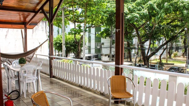 A covered outdoor seating area with wicker chairs and a white table, a white picket fence, and a view of buildings and trees. - Hospedaria Gebel LocDay (Recife)