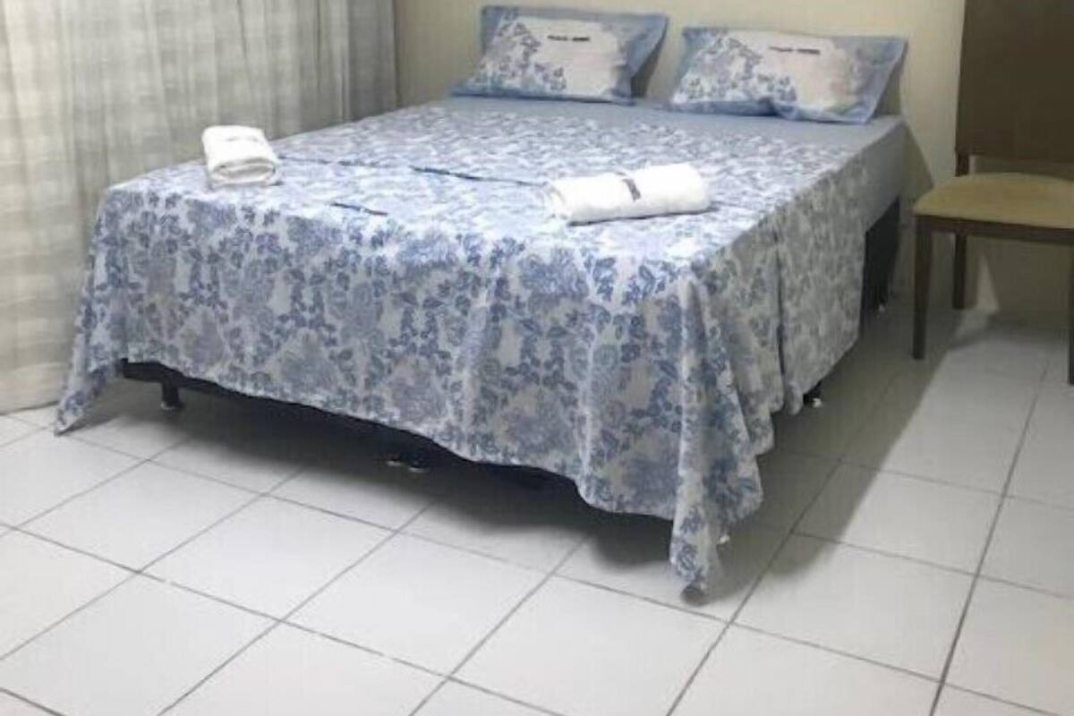 Uma cama bem arrumada com roupa de cama floral, uma pequena mesa de cabeceira e um aparelho de ar condicionado na parede.